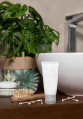 Cream tube near brush, cotton swabs and green monstera on wooden countertop in bath, mockup