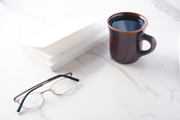 白いテーブルに置かれた一冊の本とメガネ、コーヒーの入ったマグカップで、コーヒーを飲みながら読書を楽しむイメージ

