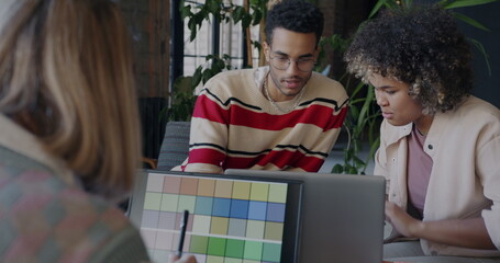 African American businessman talking to female colleague while lady designer working with laptop in creative office. Start-up and coworking concept. - 792810402