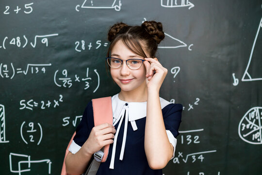 A beautiful schoolgirl adjusts her glasses on her nose while standing the school board.
