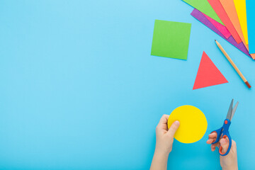 Little child hand holding scissors and cutting colorful geometric shapes from application paper on pastel blue table background. Making different forms. Point of view shot. Closeup. Top down view. - 792819428