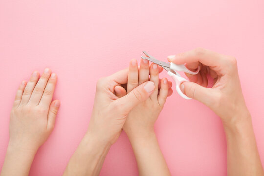Young adult mother hand holding scissors and cutting little child finger nails on light pink table background. Pastel color. Closeup. Point of view shot. Top down view.