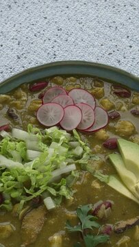 A bowl with vegan Pozole Verde dish with hominy, beans, tomatillo, and button mushrooms topped with avocado, red radish, and shredded napa cabbage. Copy space, table spin, vertical video. 