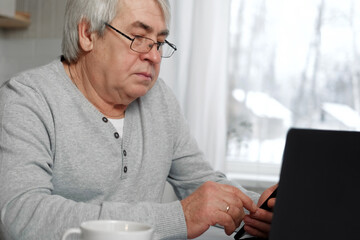 Portrait of kind Old man Sitting at Table with laptop and looking at display. Confident Mature Grey haired 60s Aged Man working at computer from home. Casual lifestyle of retired people. Senior male