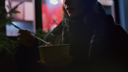 Female student donned in cozy warm clothing enjoys Asian street food walking urban streets at night