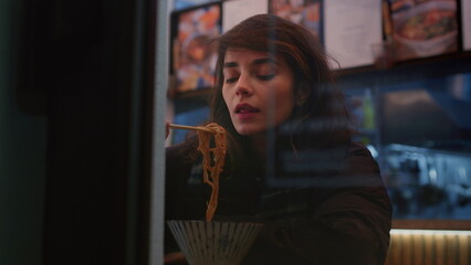 Young brunette woman eats Asian dish sitting at table in darkened premise of street Asian cafe
