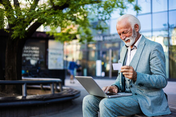 Matuure businessman sitting on concrete bench in city, he is holding a credit card and using laptop for online shopping during the day.