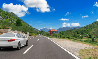 A white car drives along the highway against the backdrop of rocky mountains on a sunny day.