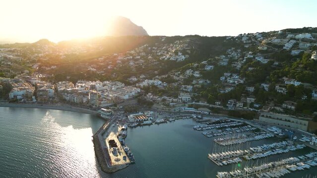 Cinematic aerial view of Xabia - Javea city at sunset. View of marina and part of the city. Sunset point. 
Sun in front of camera, setting behind mountains. Drone forward above the marina. Travel