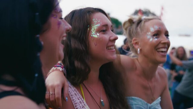 Three female friends arm in arm wearing glitter singing and dancing watching band at outdoor summer music festival - shot in slow motion 