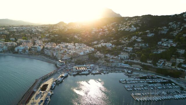 Panoramic aerial view of costal city Xabia known also as Javea. Spectacular sunset colours. Sun in front of the camera. Crone circling above the marina and panning left. Famous travel destination.
