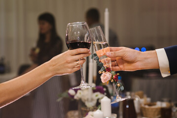 Cheers. Bride is holding glass of red wine outdoors and groom hold champagne. Wedding celebration...