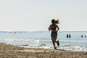 Back view of a young woman with a ponytail jogging along the seashore on a sandy beach wearing...