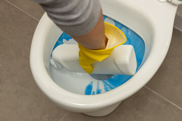 Close up of hand wearing a yellow protective glove and applying a blue liquid toilet cleaner to...