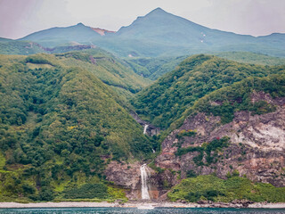 A distant view on a late summer afternoon from a sightseeing cruiser over the famous landmarks of Kamuiwakka (Water of the Gods) Onsen Falls and Mt. Iou in Shiretoko Peninsula, East Hokkaido, Japan.