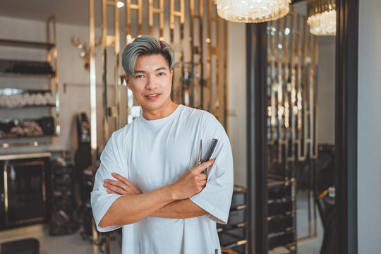 Happy asian professional Hairdresser or hair stylist man standing confidence with arm crossed with smile and holding hairdressing equipment while looking at camera in beauty salon and barber shop