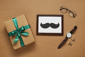 Gift box, frame with paper mustache and men accessories on brown background, flat lay. Father's day...