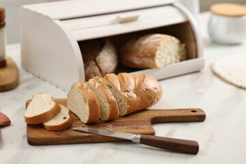 Wooden bread basket with freshly baked loaves and knife on white marble table in kitchen