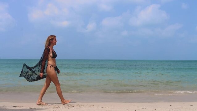 Side shot of young caucasian woman in bikini and penuar walking through the sea beach. Slow motion and wide shot.