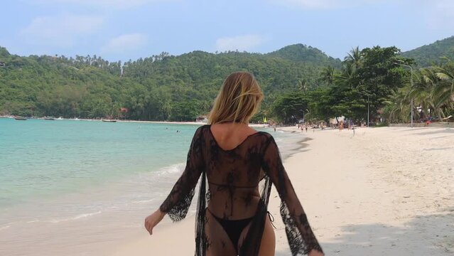 Back view of young carefree happy caucasian woman in bikini and penuar walking through the sea beach. Slow motion and wide shot.