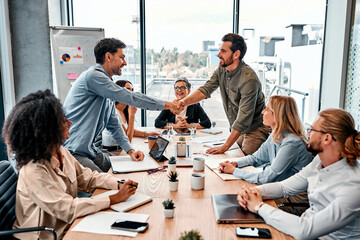 Deal approving. Millennial caucasian workers coming to agreement and shaking hands by desktop during meeting. Corporate managers gathering in conference room with wooden table and whiteboard.