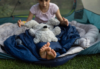 barefoot child sits in a tent, covered with a sleeping bag, and plays with favorite teddy bear....