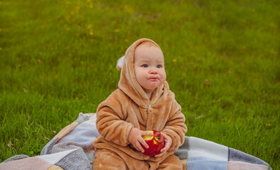 A cute little baby girl in a warm jumpsuit is sitting on a plaid on a green lawn with a red apple