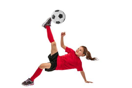 Portrait of girl, child in red uniform training football techniques against transparent background. Kicking ball in jump. Sportive and active kid. Concept of action, team sport game, energy, vitality.