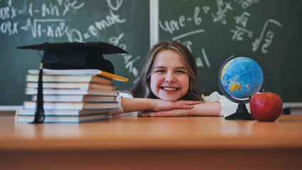 Adorable school girl posing at her desk against a background of blackboard, books, globe and graduation cap.