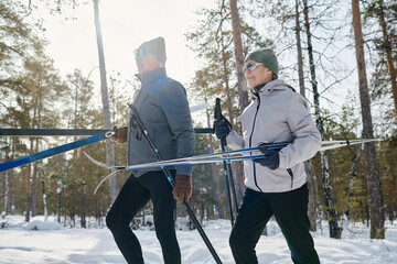 Senior Caucasian man and woman in winter sportswear holding skis and poles walking along forest park, medium long shot