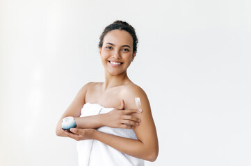 Amazing hispanic or brazilian young woman standing on white background wrapped in white towel, applying nourishing cream on body, looks at camera, smiles. Caring of a body. Skin care concept