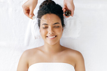 Top view of a beautiful well-groomed smiling hispanic or brazilian woman, wrapped in a white towel, lying during a procedure with a cosmetologist which applying on her forehead hyaluronic serum