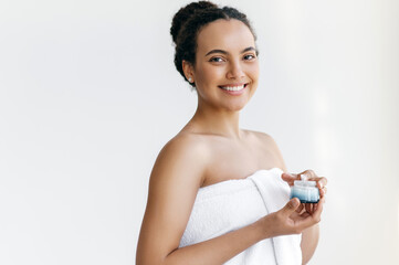 Beautiful cute hispanic or brazilian young woman, wrapped in white towel, holding nourishing body cream in a hand, preparing to apply to skin after shower, looks at camera, smiling. Skincare routine