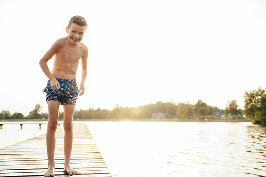 Cunningly laughing frozen teenage boy stands in wet swimming trunks on a wooden bridge before jumping into a lake at sunset in the summer.