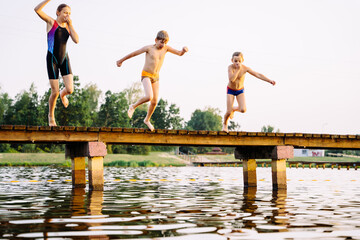 Teen girl in swimsuit and two smaller boys jumping from a bridge into a lake at sunset.