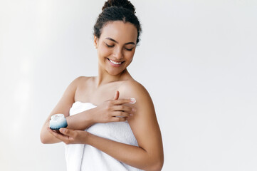 Caring of a body. Happy beautiful hispanic or brazilian young woman standing on a white background wrapped in white towel, gently applying moisturizing cream on body, smiling