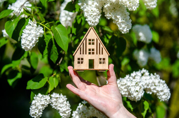 The girl holds the house symbol against the background of blossoming white lilac
