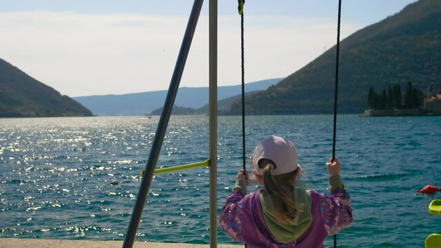 Close-up of a little girl riding on a swing on the seashore and admiring the beautiful view. Back view. Perast, Montenegro.