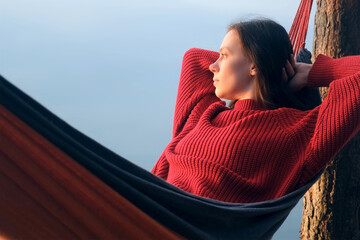 Woman relaxing admiring lake lying in hammock in forest at sunset on nature. Resting young female in red sweater enjoying time in camping in woodland. Tourist traveler, camper outdoors.