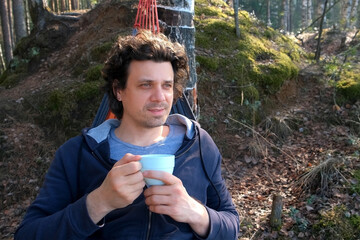 Portrait curly man relaxing lying in hammock drinking tea in forest on nature. Resting relaxed brunet male enjoying time in camping in woodland. Tourist, traveler, camper contemplating outdoors.