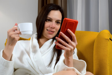 Portrait of brunette woman in bathrobe is drinking tea, laughing and browsing smartphone in hotel in evening sitting in yellow cozy armchair. She is resting smiling looking at phone screen on weekend.