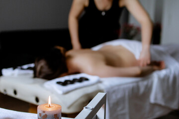 Burning candle in the foreground. Woman enjoying hot stone massage at spa salon. Professional masseur making stone therapy. Relaxing and ease tense muscles and damaged soft tissues throughout body.