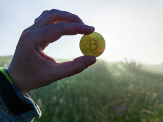 Caucasian hand holding a Bitcoin shiner on a morning grassy meadow background