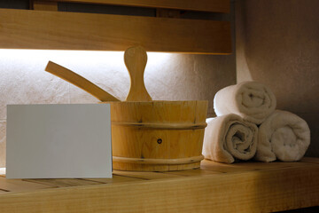 Close-up of a blank white paper card, sauna bucket with ladle and a stack of three rolled white towels on wooden bench in sauna lit by back intimate light. Wellness and well-being concept