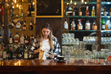 Waitress standing in bar and wiping and polishing beer glass. Bartender woman working in bar counter, preparing and cleaning, work before guests come, surrounded by drinks.