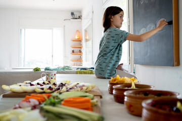 girl drawing on blackboard sitting at a table, in the foreground with vegetables and healthy buffet...