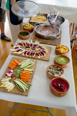 zenithal view of table with healthy food, wooden board with raw vegetables cut into strips and endive leaves to go with hummus, several bowl with chickpea hummus, in the background hot pot of beans