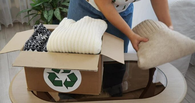 A woman activist collects old clothes for recycling or selling in a cardboard box. Female hands folded old clothes into a cardboard box on the table. Eco-friendly, recycling, second hand.
