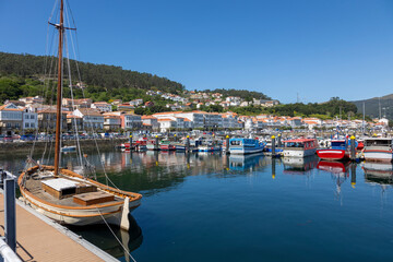 Fototapeta na wymiar A serene marina of Muros in Spain with various boats docked in calm waters, with quaint hillside houses and clear blue sky in the background
