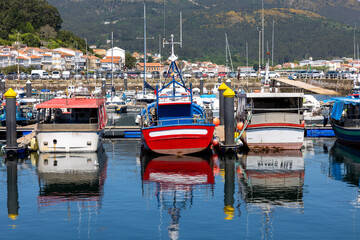 A serene marina of Muros in Spain with various boats docked in calm waters, with quaint hillside houses and clear blue sky in the background - obrazy, fototapety, plakaty